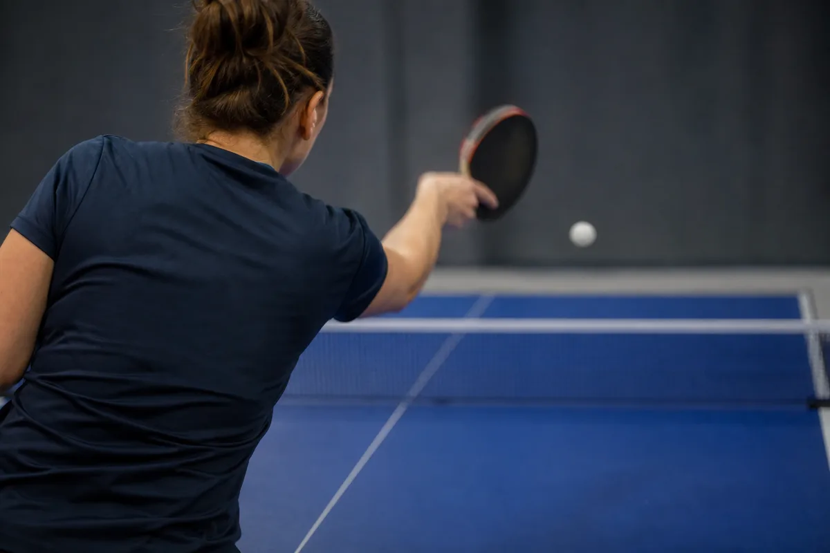 Joueuse de dos répétant un geste de revers devant une table de tennis de table dans un gymnase